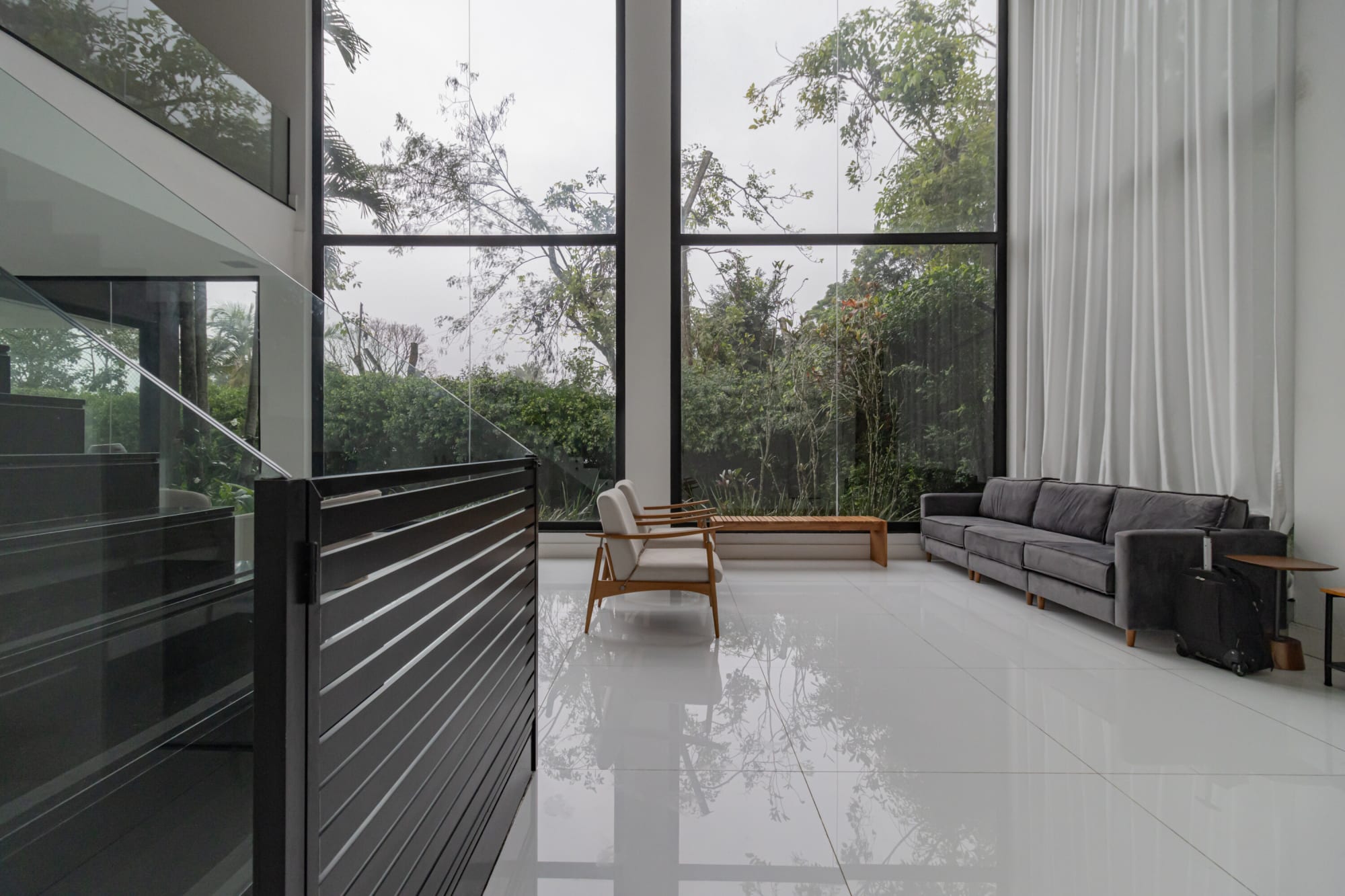 Double height glass living room with grey sofa, sheer curtains and forest view