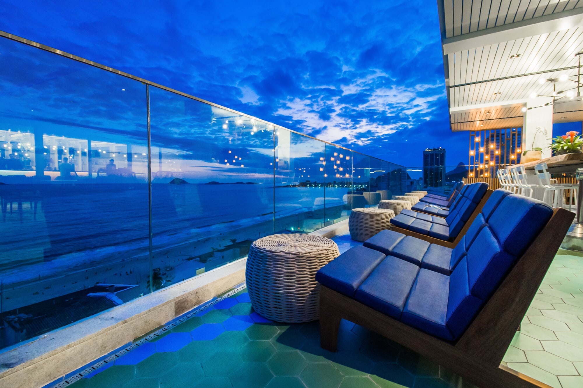 Rooftop deck at night with blue cushioned loungers behind a glass rail over the bay