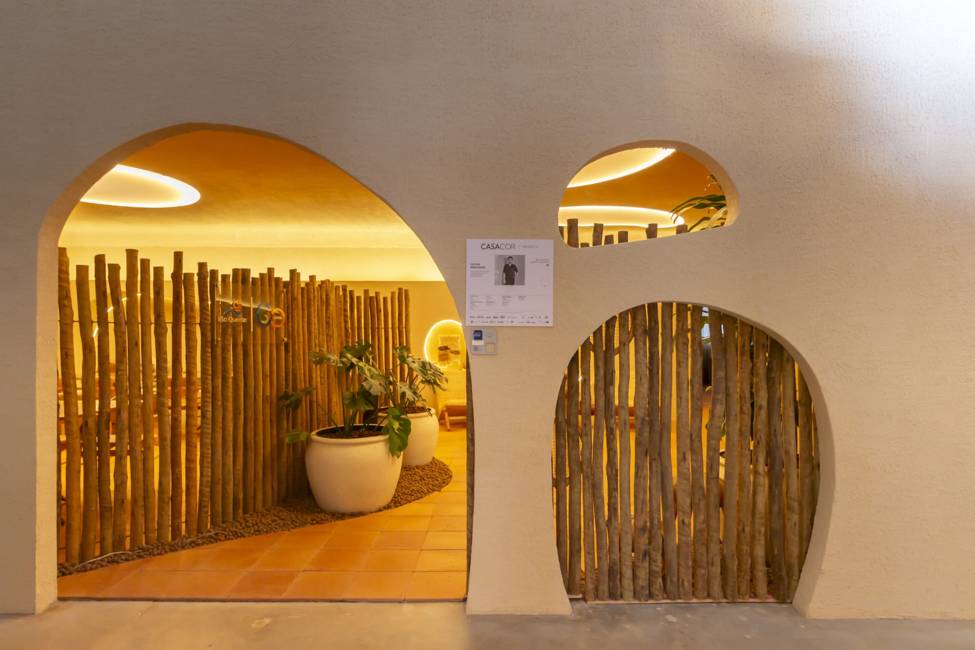 Arched plaster doorways framing timber pole screens and a potted plant in the CASACOR resort lounge