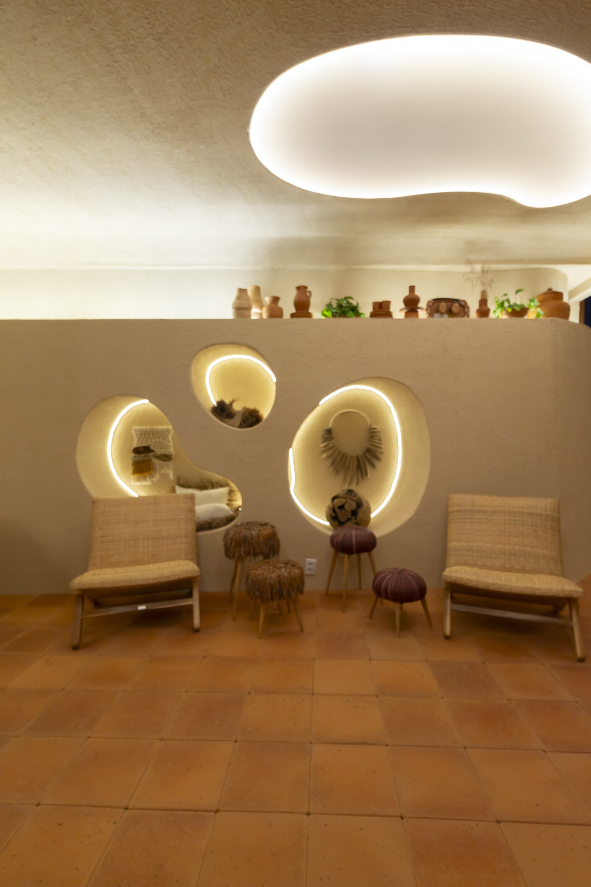 Plaster wall with glowing oval niches above woven lounge chairs and small stools on terracotta tile