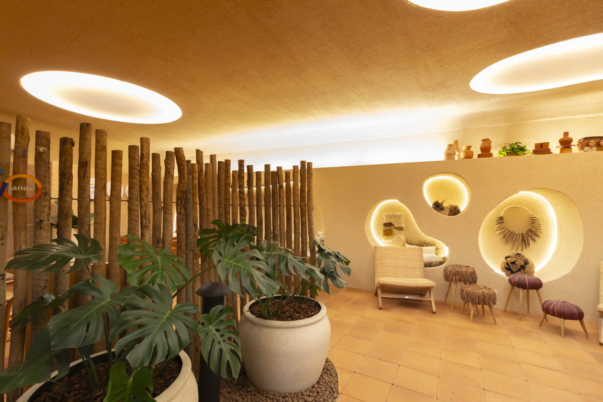 Monstera in a white planter before timber poles and lit plaster niches in a terracotta floored lounge