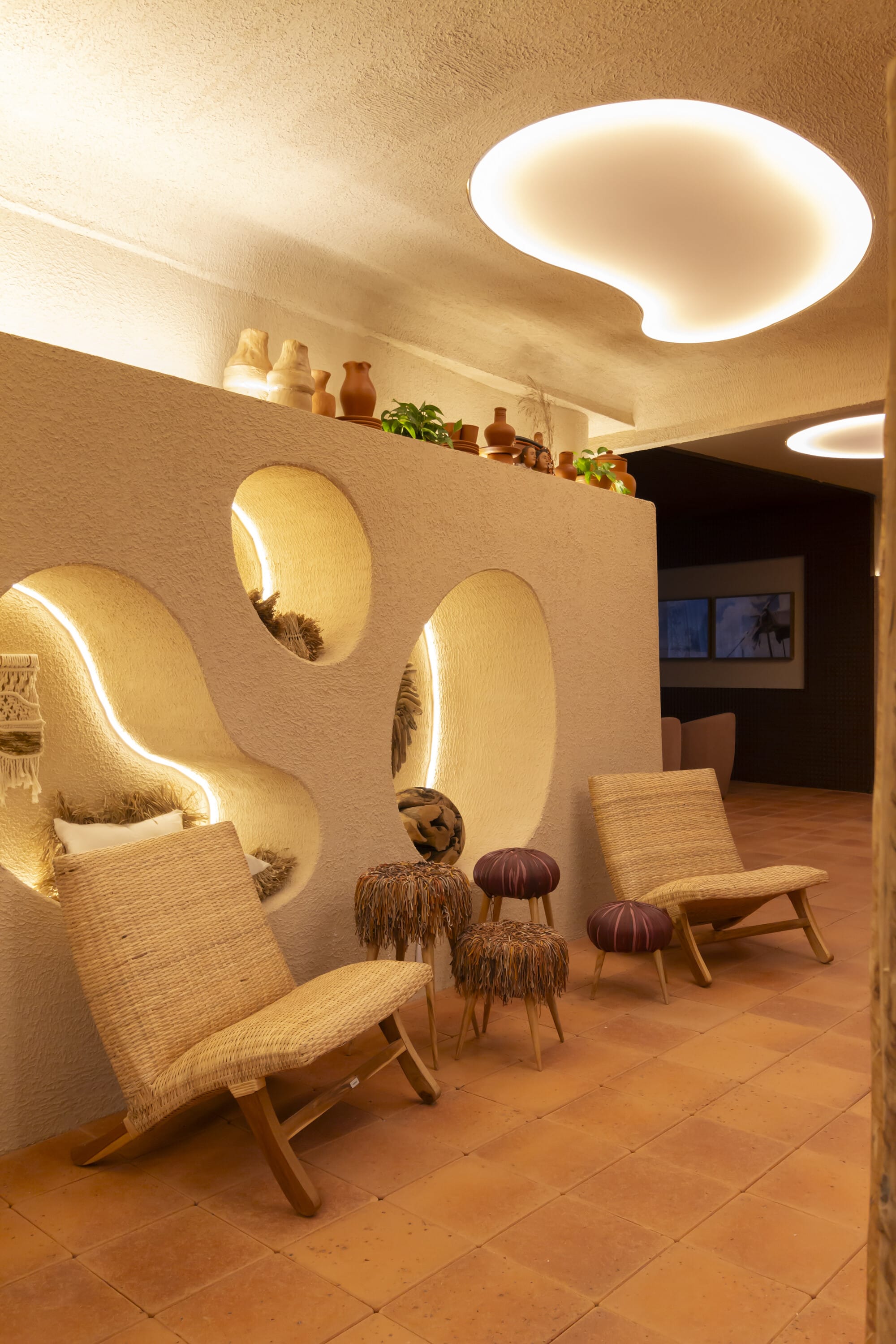 Woven lounge chair beside lit oval niches in a curved plaster wall with pottery on top
