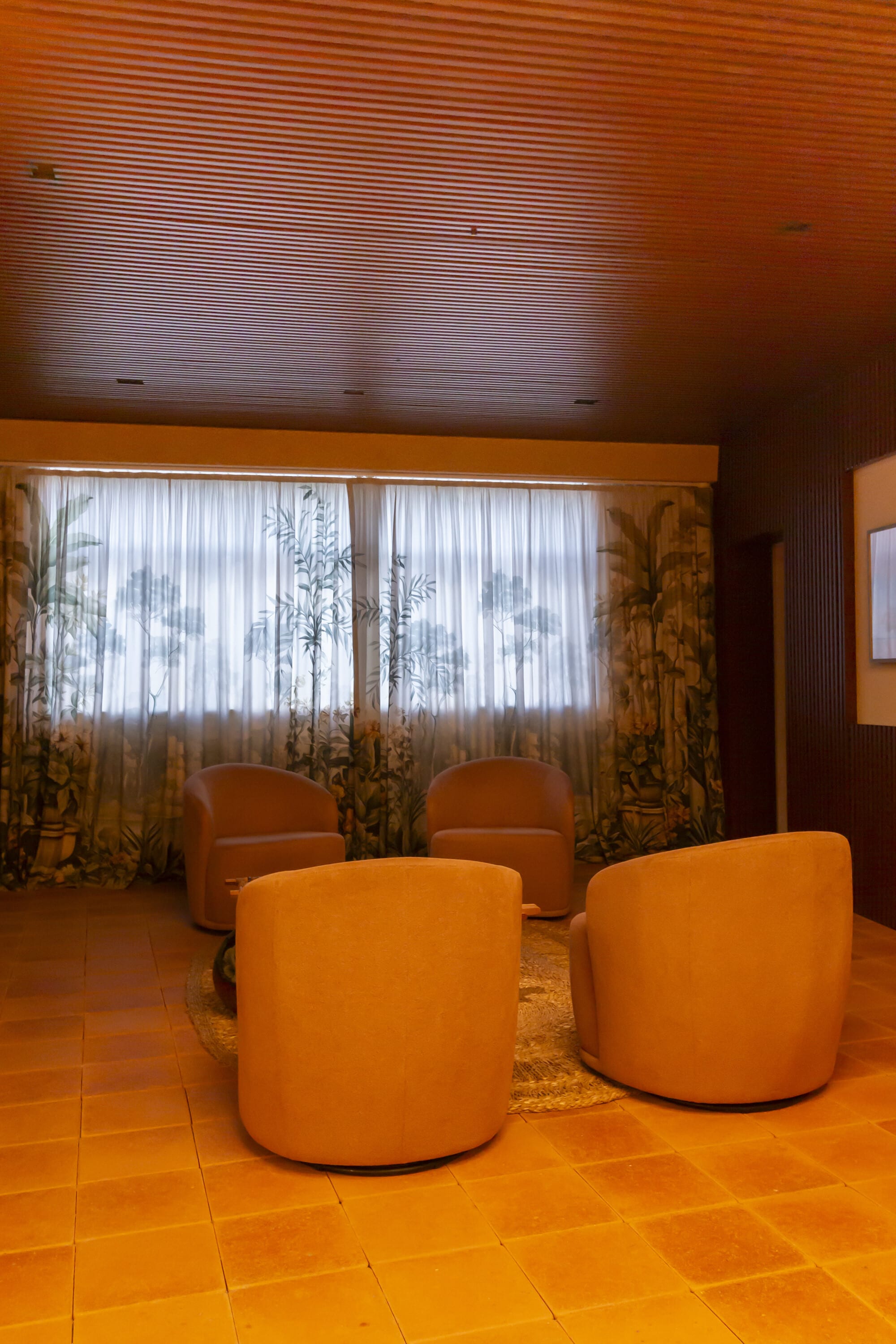 Four orange swivel chairs before botanical print curtains under a slatted timber ceiling