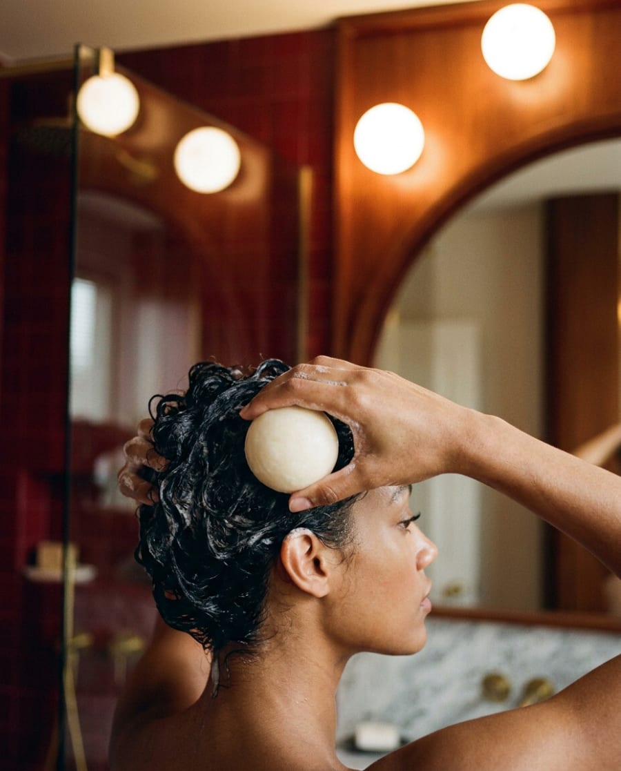 Round cream shampoo bar lathered into curls in an oxblood bathroom with globe lights