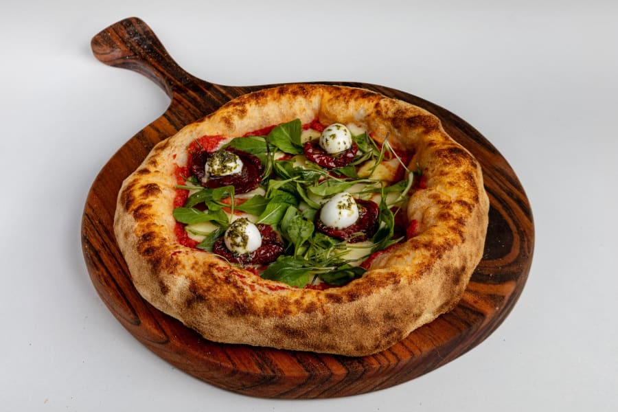 Caprese pizza with burrata, rocket and sun dried tomato on a wooden peel against grey