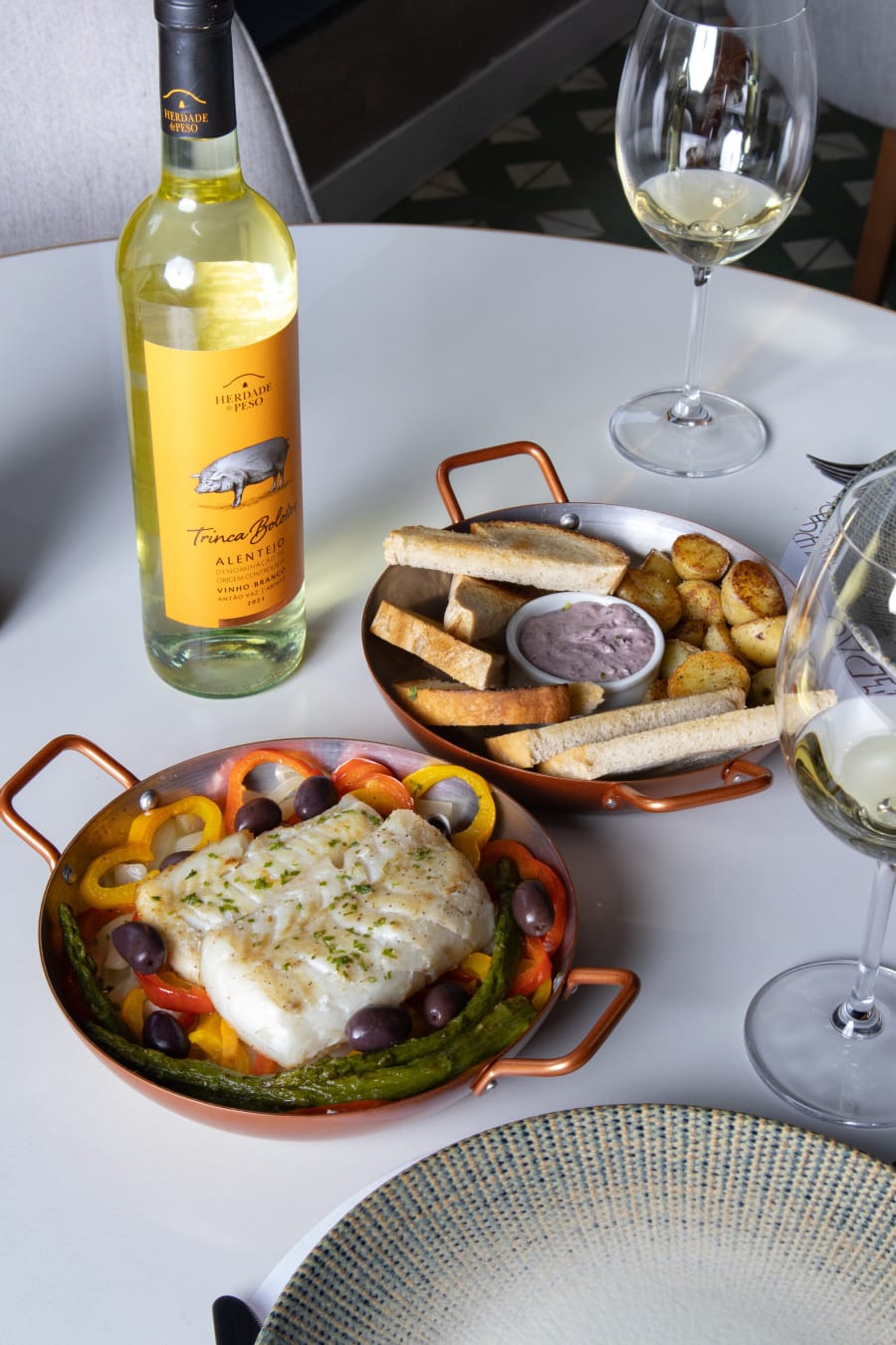 Baked cod in a copper pan with olives, bread sticks and white wine on a round marble table