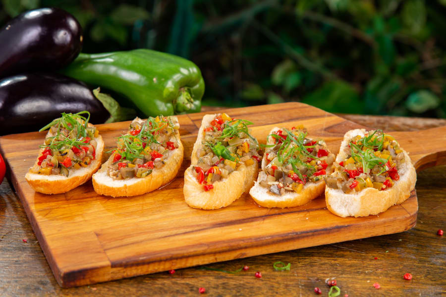 Five bruschetta slices with diced tomato and herbs on a wooden board, aubergines and peppers behind