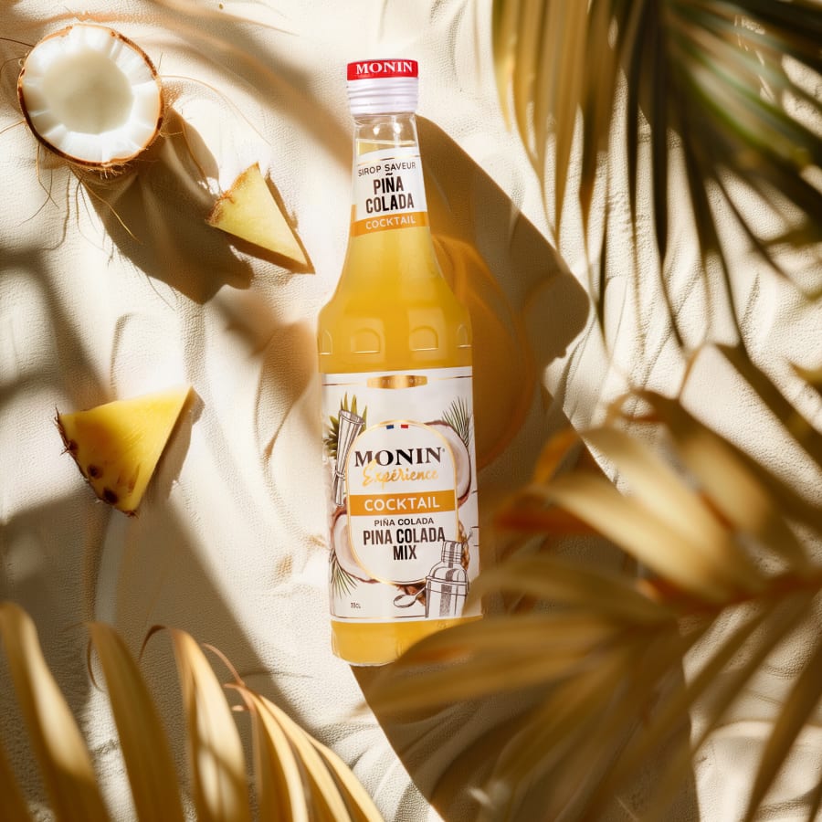 Monin Piña Colada mix bottle floating with coconut and pineapple pieces in palm leaf shadow