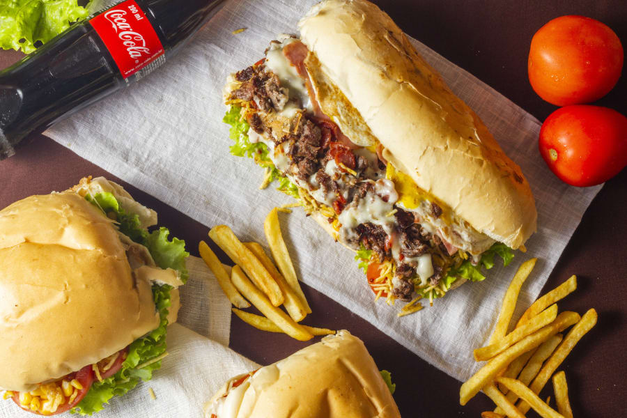Steak sandwich and burgers on paper with fries, tomatoes and a Coca Cola bottle on dark wood