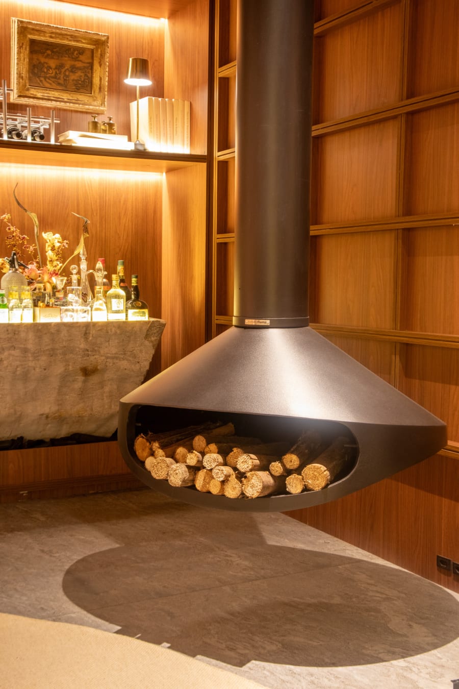 Black hanging fireplace with stacked logs before a lit stone bar in walnut panelling