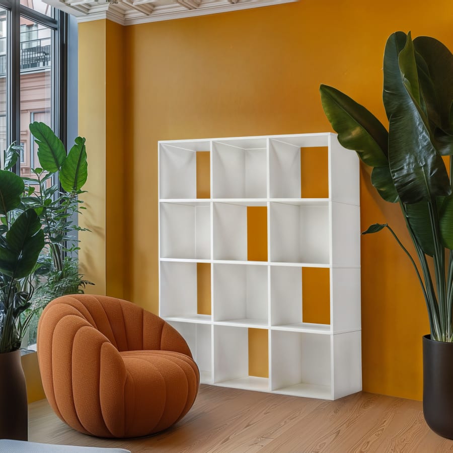 A white Movêu cube bookcase against a mustard wall beside an orange tufted armchair