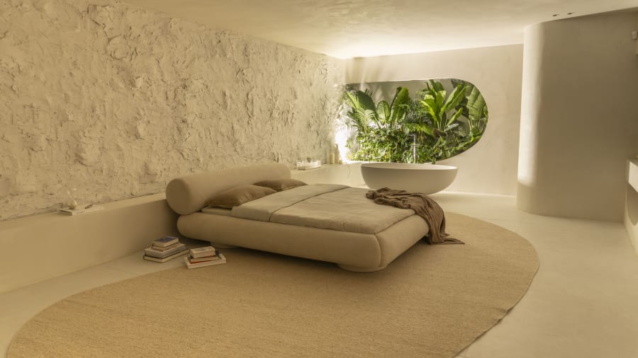 Low upholstered bed on an oval rug in a lime plaster room with a planted window