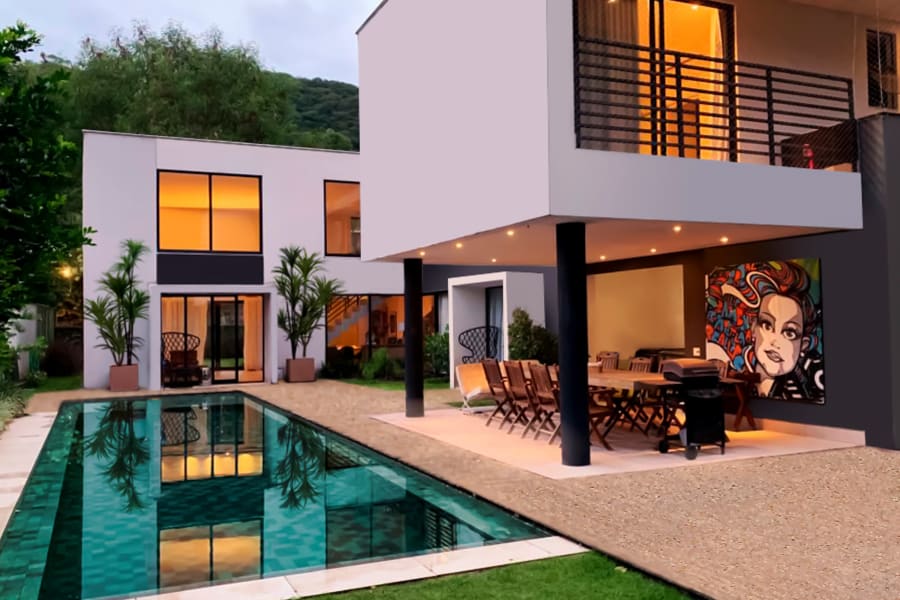 White cubic house at dusk with lit windows, covered terrace and long green tiled pool