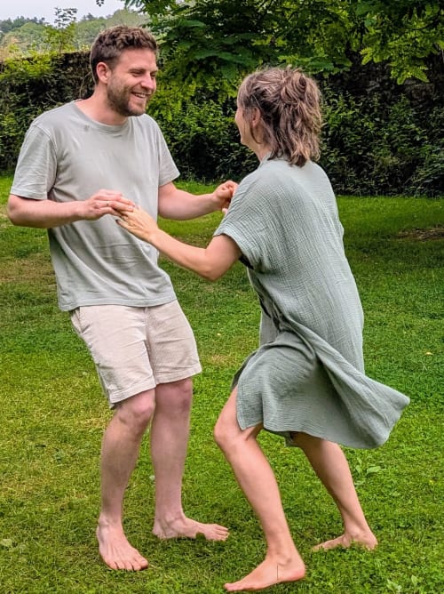 Zwei barfüßige Personen stehen auf einer Wiese und halten sich spielerisch an den Händen. Eine trägt ein graues T-Shirt und beige Shorts, die andere ein grünes, locker fallendes Kleid. Im Hintergrund sind Bäume und Büsche zu sehen.