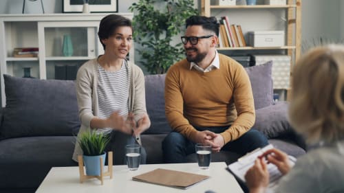 Zwei Personen sitzen in einem Beratungsgespräch auf einem Sofa; eine dritte Person macht sich Notizen.