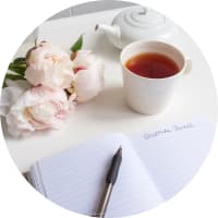 Aufgeschlagenes Notizbuch mit Stift, daneben eine Tasse Tee und Blumen