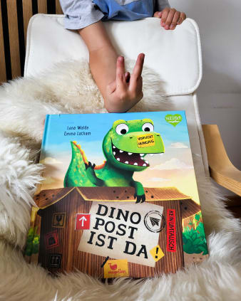 Produktbild - Dino-Post ist da! - Buch mit Cover auf Kuschelfell und Stuhl, dahinter Kinderhand