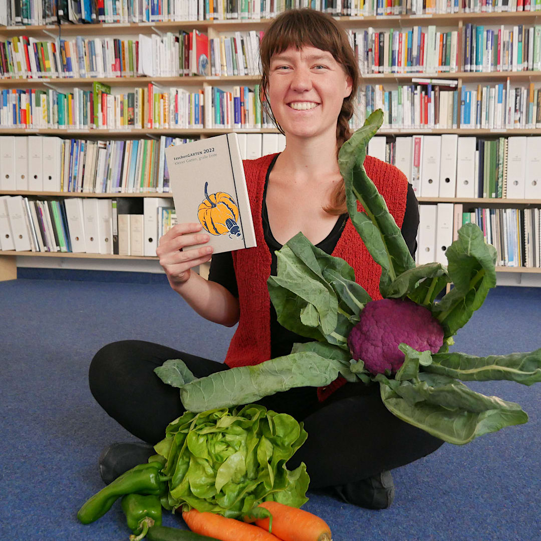 oekom-Mitarbeiterin Hannah sitzt auf dem Boden und hält den TaschenGarten und verschiedenes Gemüse