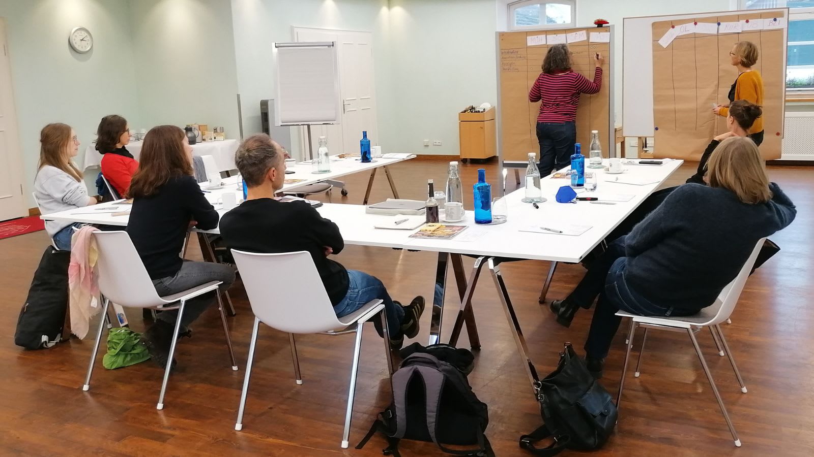 Konferenzraum mit Tischen un Hufeisenform, an denen Menschen sitzen. Im Hintergrund arbeiten 2 Menschen an Pinnwänden