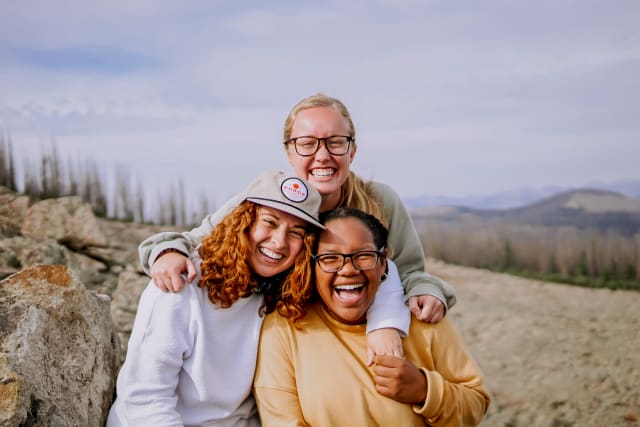 Three girl Forge students smiling and hugging each other on top of a mountain