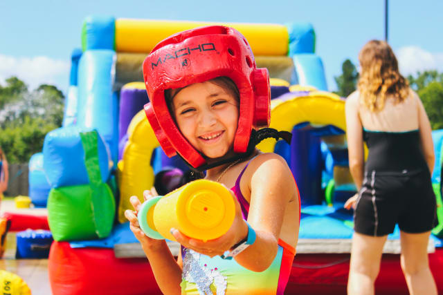 A girl wearing a soft helmet points a water toy at the camera in front of a giant, colorful inflatable