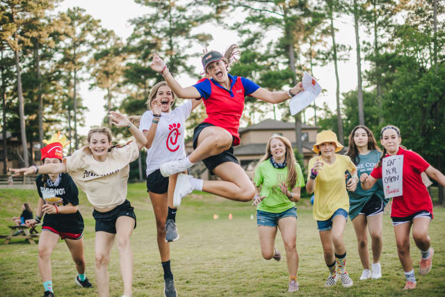Eight female campers, including one counselor jumping high in the center, are running and cheering on a grassy field in front of cabins at Timbers at Pine Cove.
