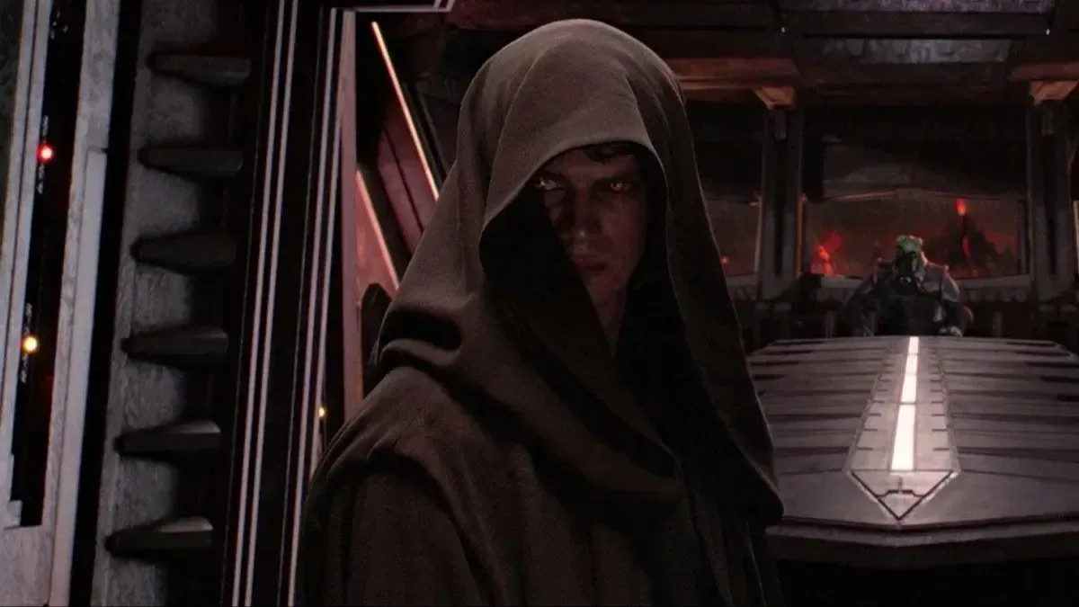 Descrição de imagem: Cena interna do filme Star Wars. Anakin está com um capuz marrom escuro tampando quase todo o rosto, deixando visível o lado esquerdo da face e seus olhos brilhando em um marrom amarelado pouco natural. No fundo uma longa mesa de metal, e mais ao fundo, pela janela, uma paisagem vermelha e montanhosa.<br>