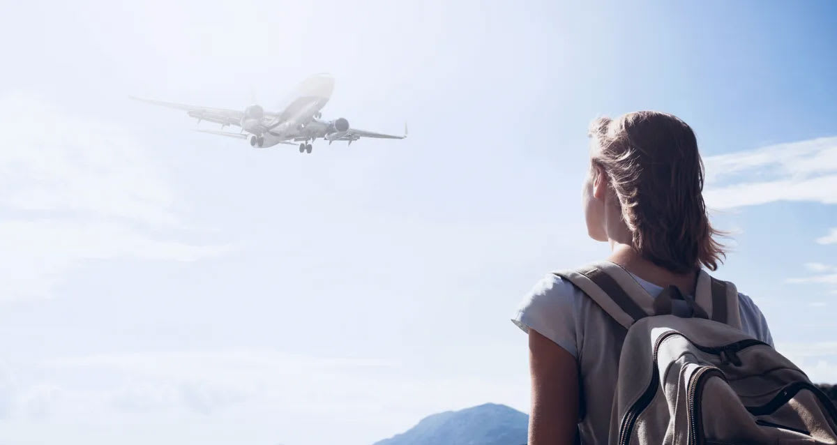 A traveler wearing a backpack looking into the clear blue sky at an airplane