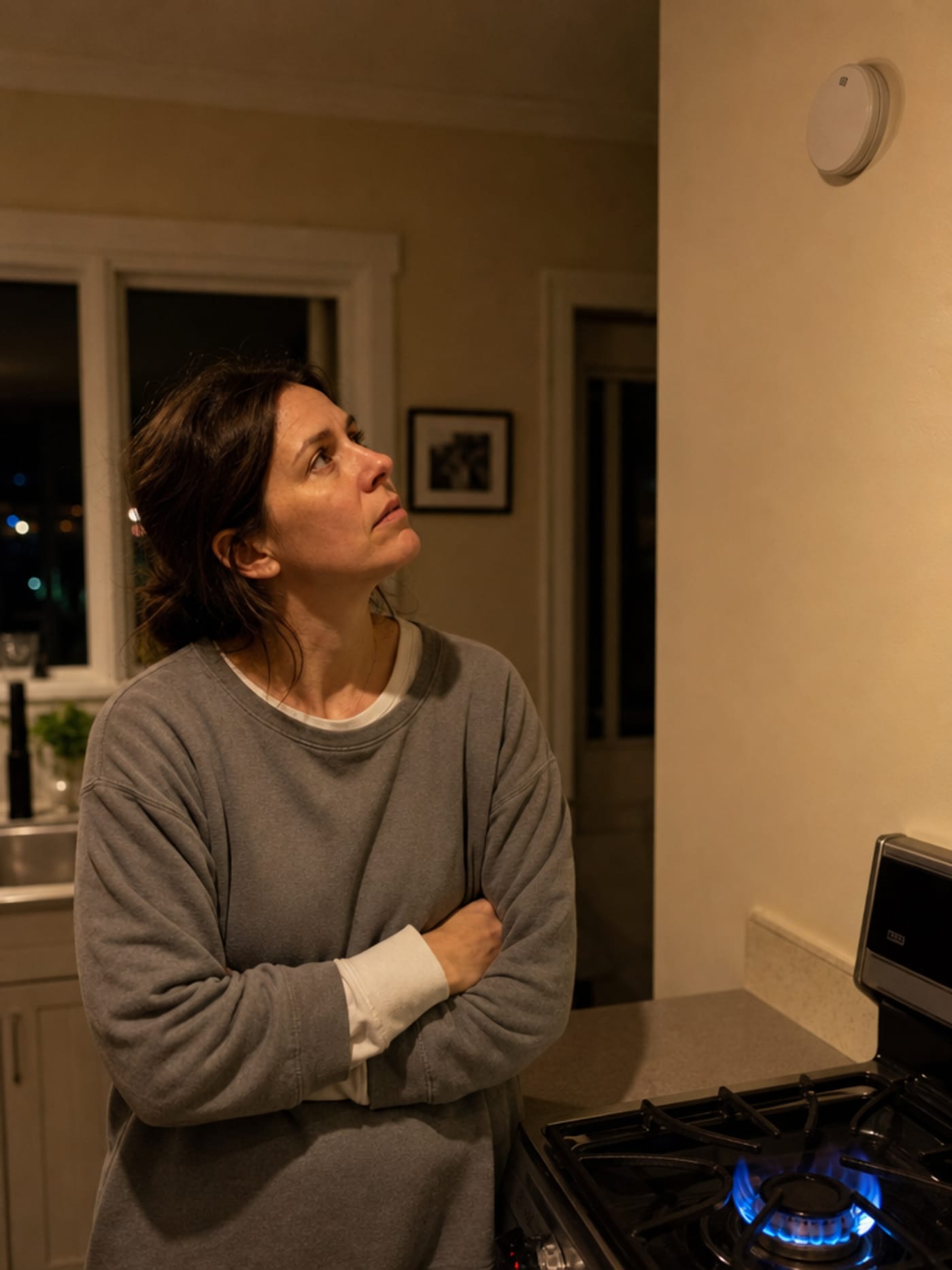 A woman standing in a kitchen at night looking up at a silent CO alarm puck on the wall, a gas stove burner lit beside her, her expression quietly uncertain