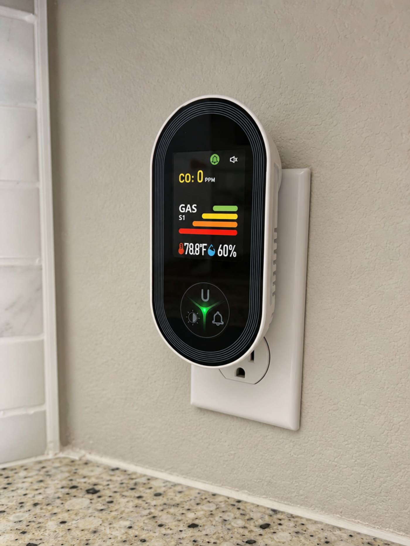 The VIGILOX monitor plugged directly into a US kitchen wall outlet, its color IPS display showing a live CO reading of 0 PPM alongside temperature and humidity figures
