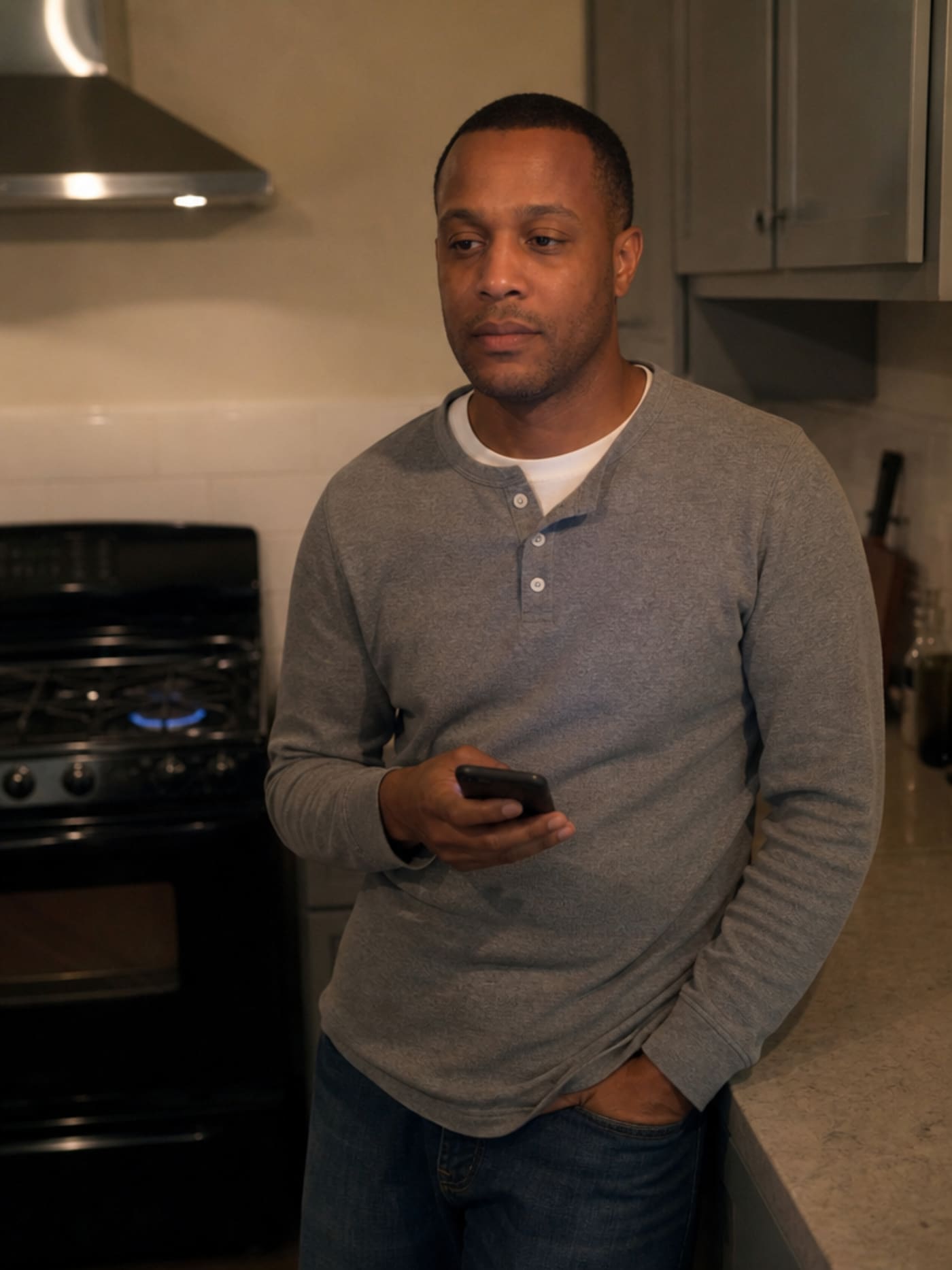 A man of 41, close-cropped dark hair, wearing a grey henley, leaning against a kitchen counter with his phone in hand — expression neutral and unaware, a gas stove burner glowing low in the background. Nothing looks wrong.