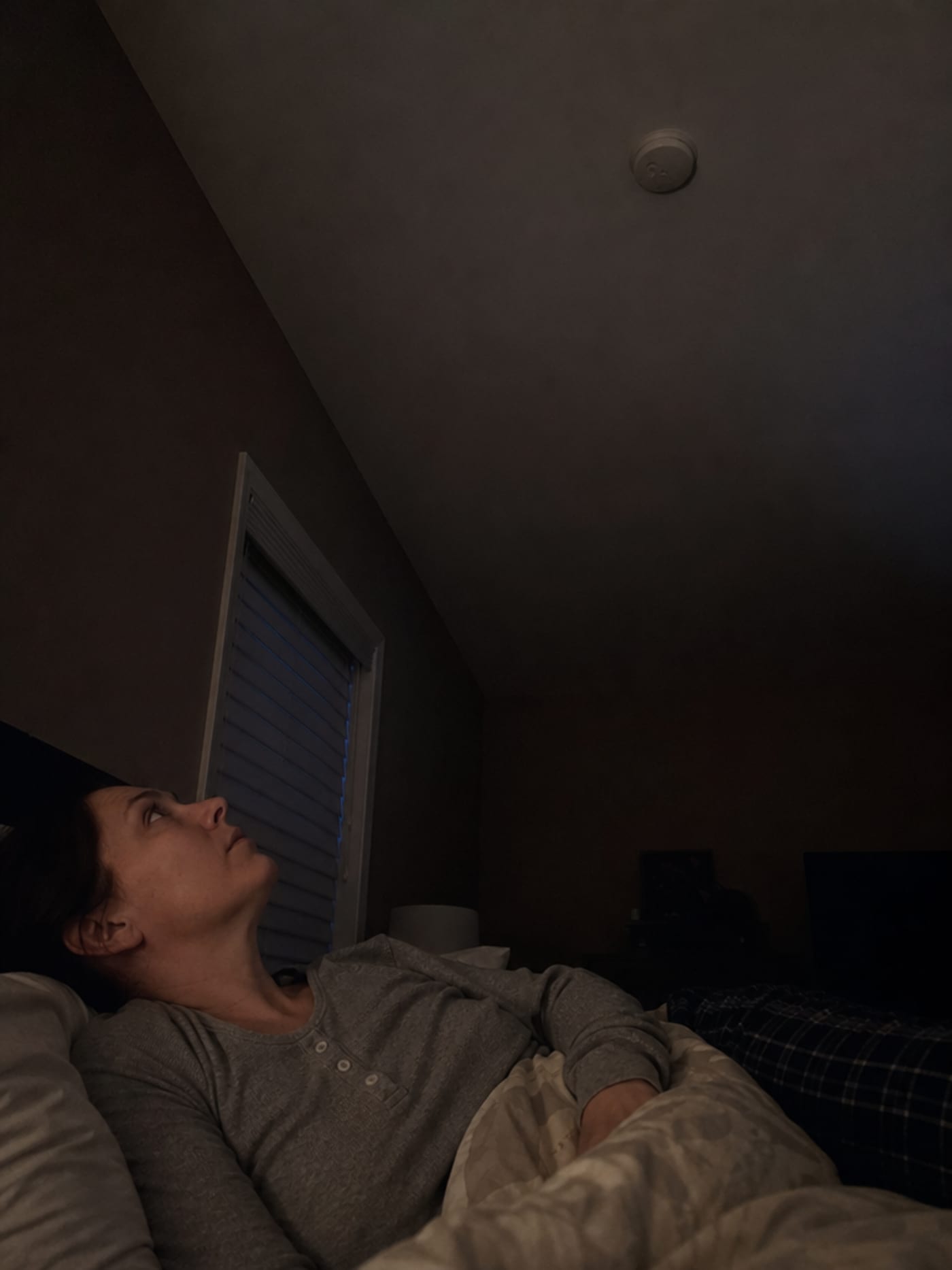 A woman lying awake in a darkened bedroom, eyes open toward the ceiling, where a silent CO detector hangs dark and unlit — the look of someone waiting for information they do not have