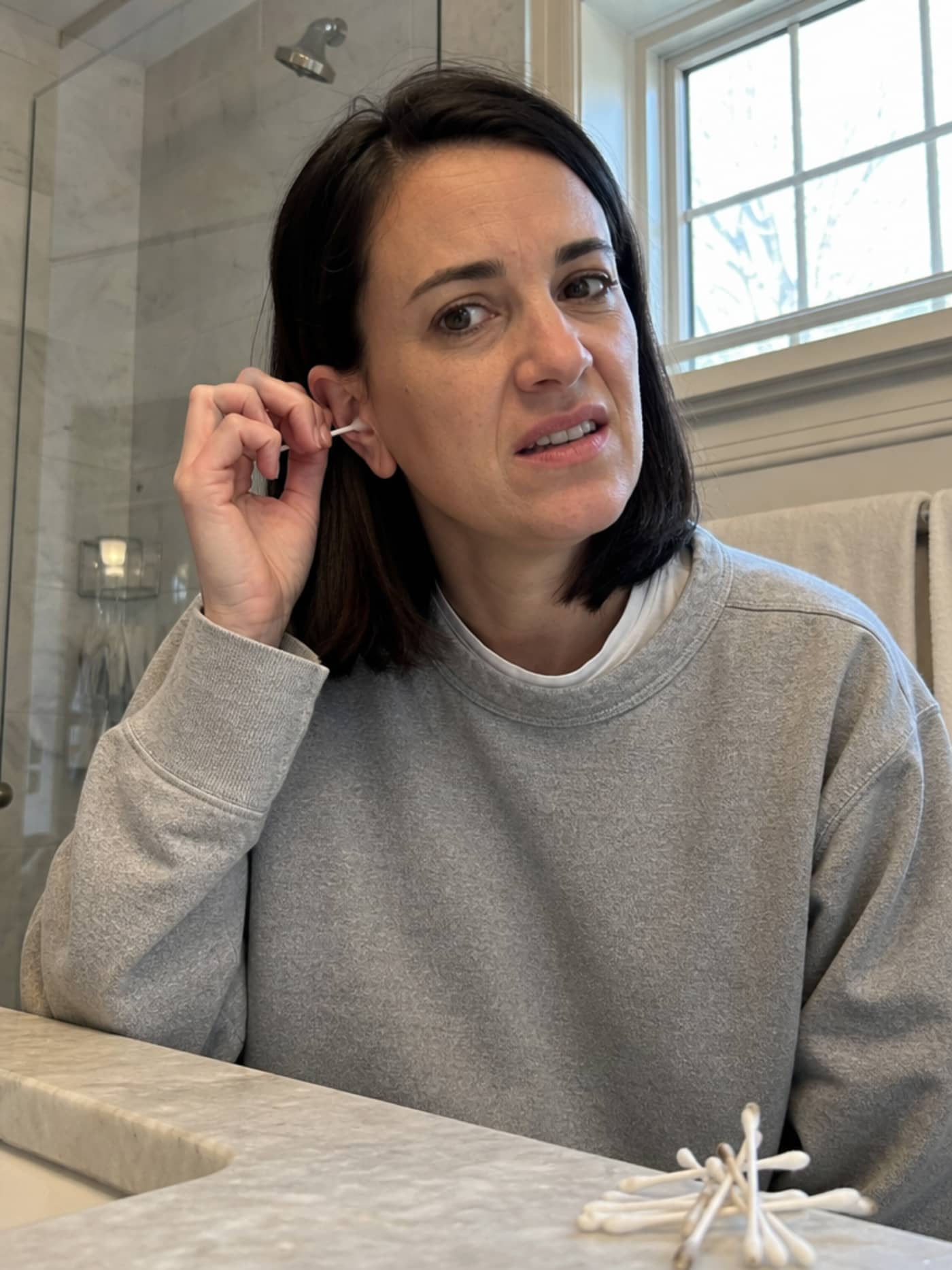 Woman with cotton swab near her ear, expression of frustrated hesitation, small pile of used swabs on the bathroom counter