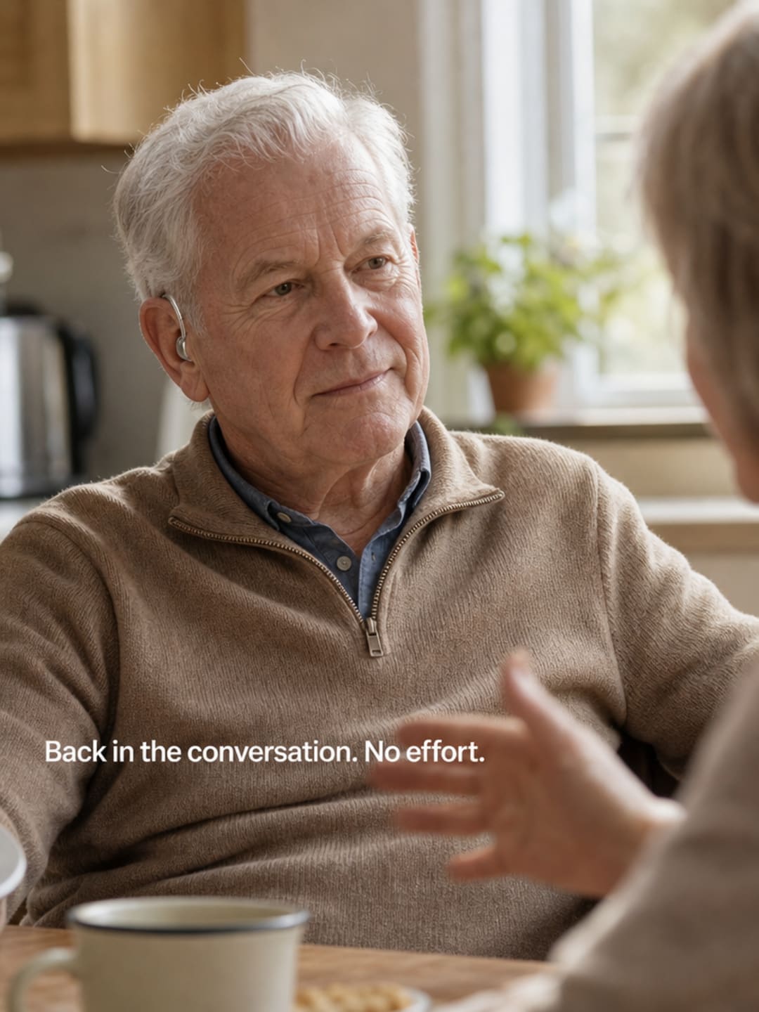 Older adult seated at dining table in relaxed easy conversation, hearing device subtly visible behind ear, warm window light