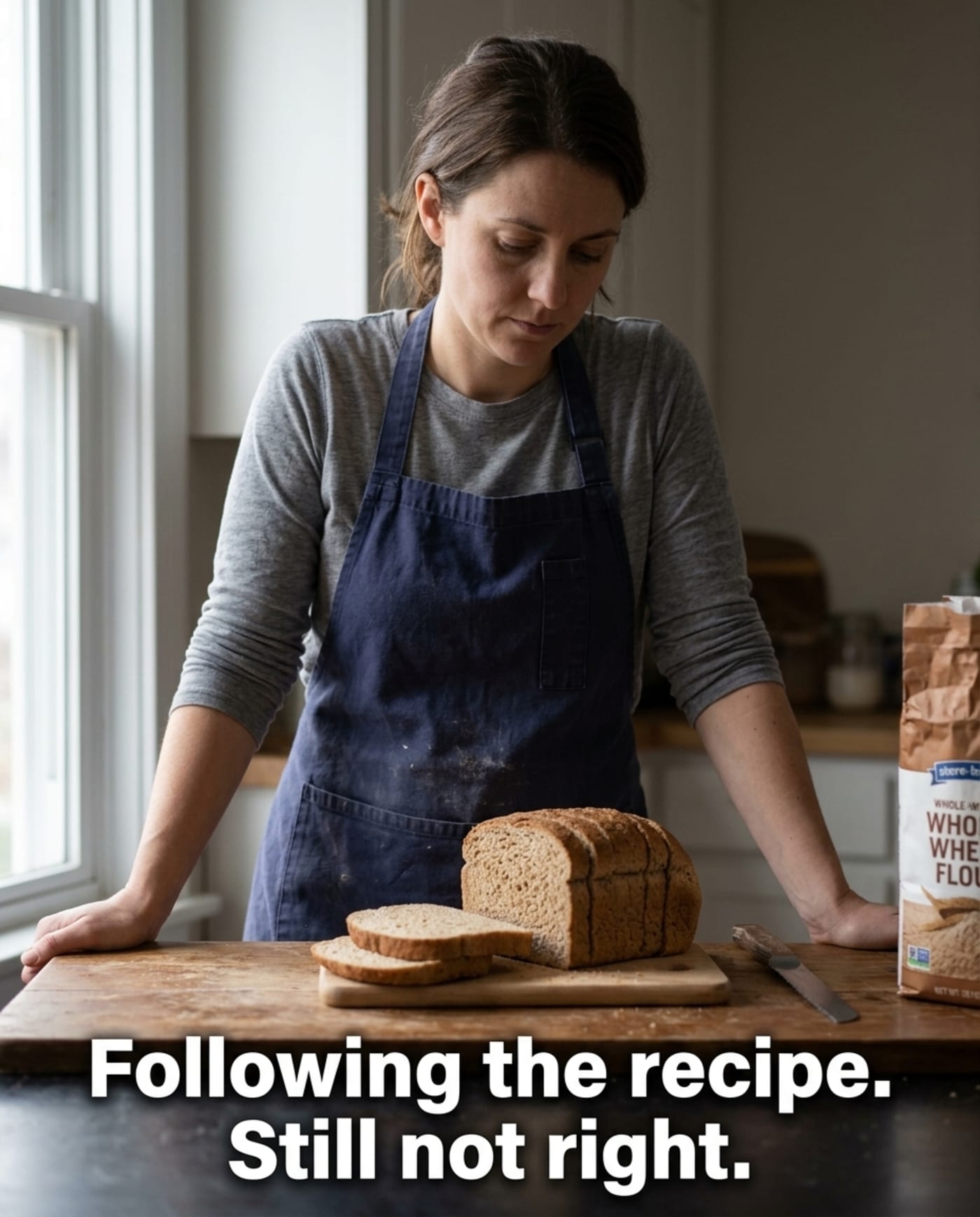 A woman in a kitchen looking down at a dense, flat loaf of bread she has just sliced, a bag of store-brand whole-wheat flour visible in the background