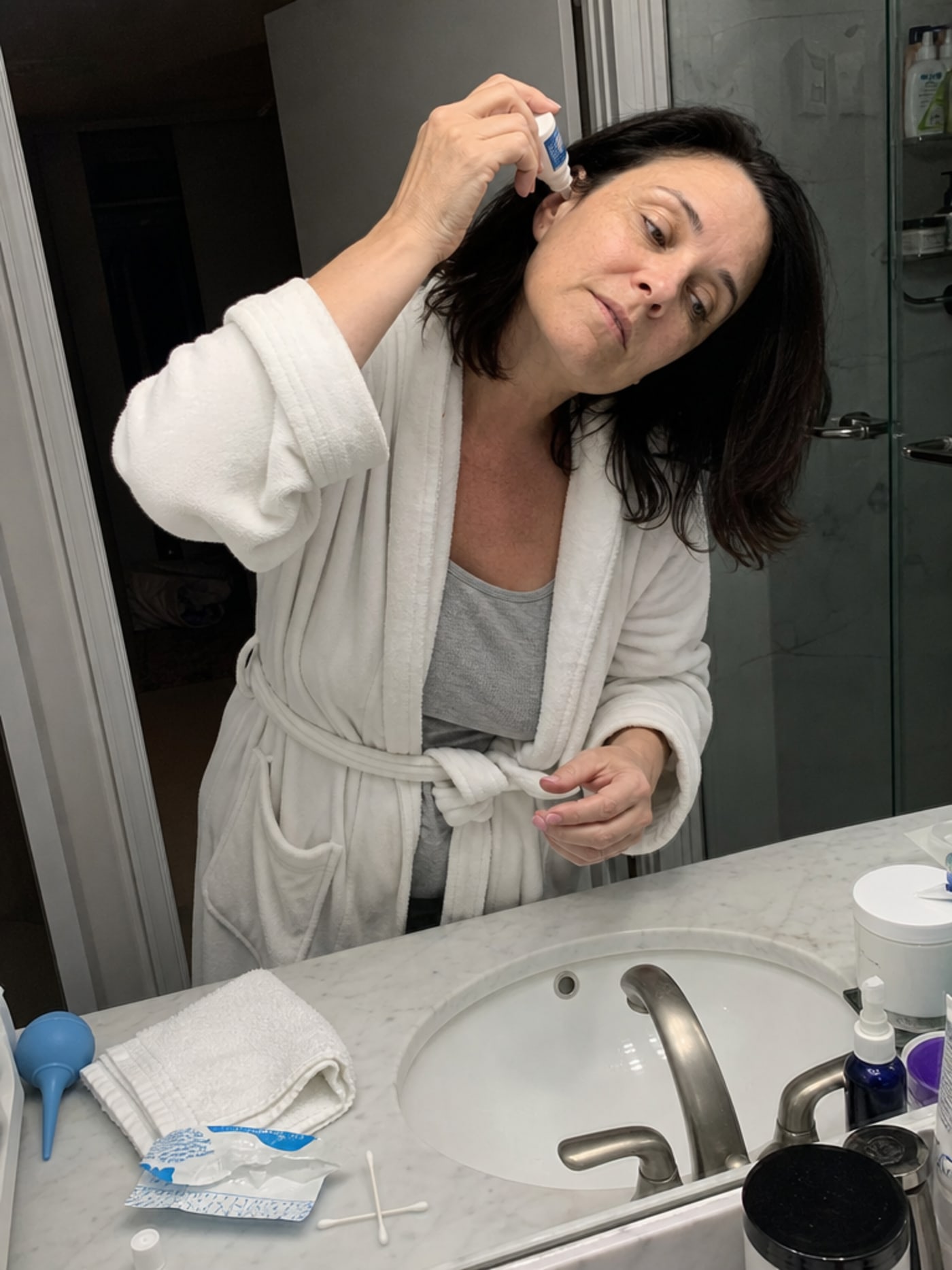 A woman of 44 with dark shoulder-length hair in a white cotton robe tilts her head over a bathroom sink holding ear drops, while a cluttered counter shows cotton swabs, a folded towel, and a bulb syringe — the resigned look of someone going through a routine she knows might not fully work