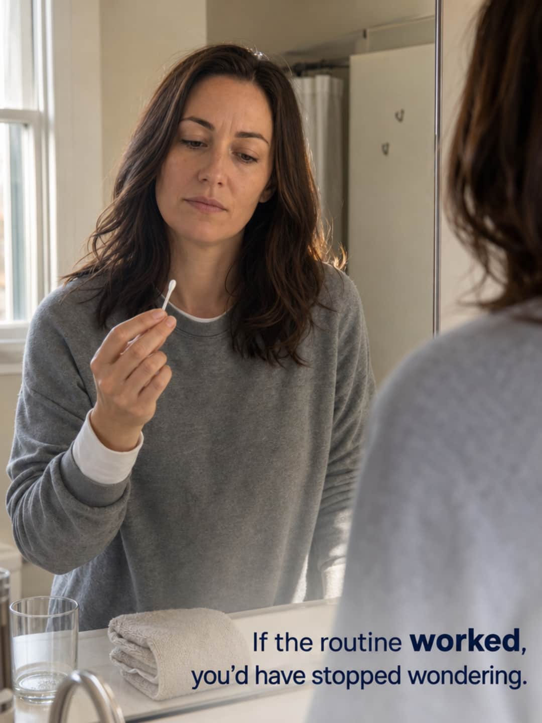 Woman at bathroom mirror pausing to examine a cotton swab with mild, untheatrical weariness — questioning whether the routine is working.