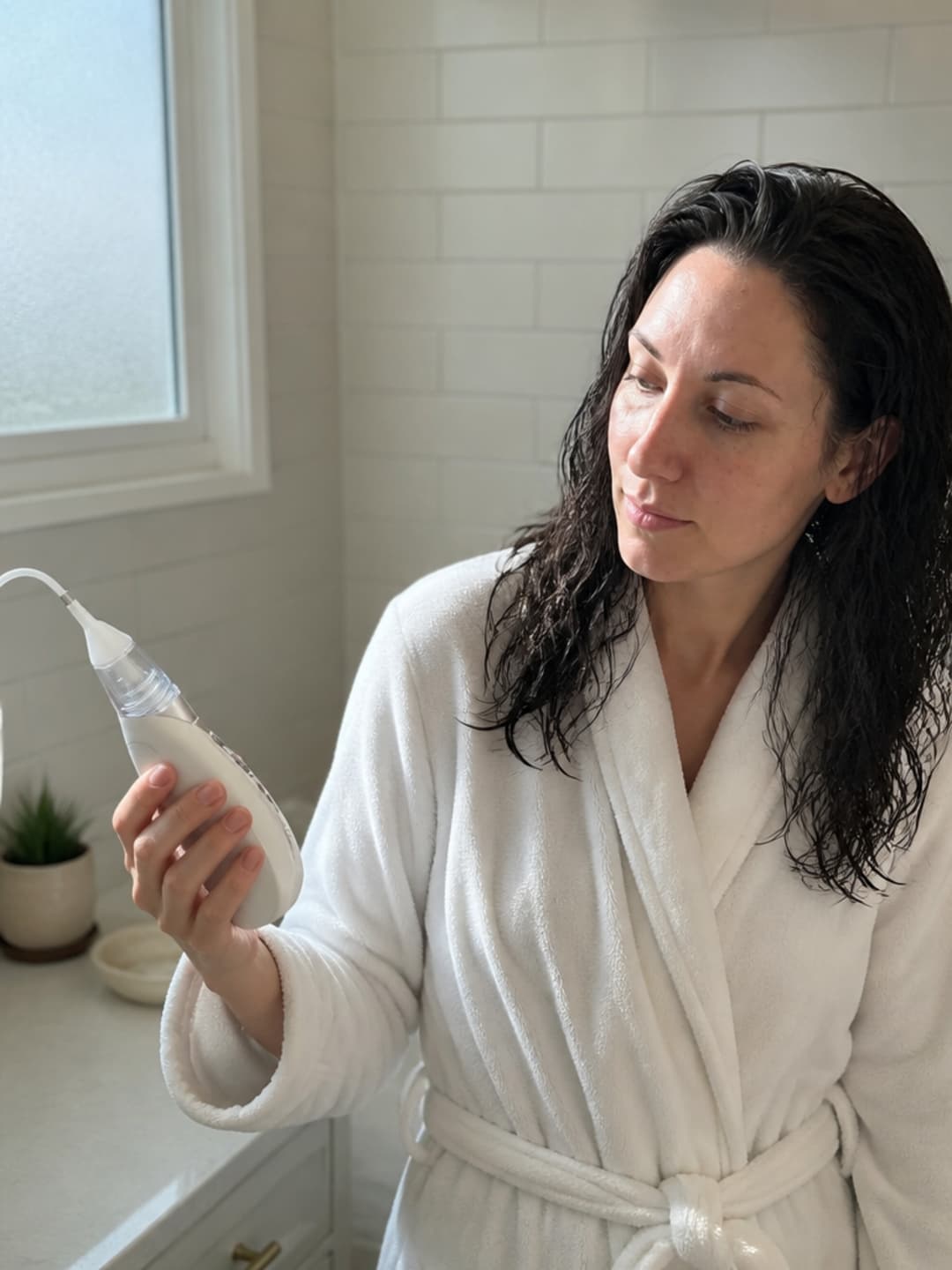 A woman in her mid-thirties in a warm bathroom holding the Vacurio device, examining it with quiet curiosity