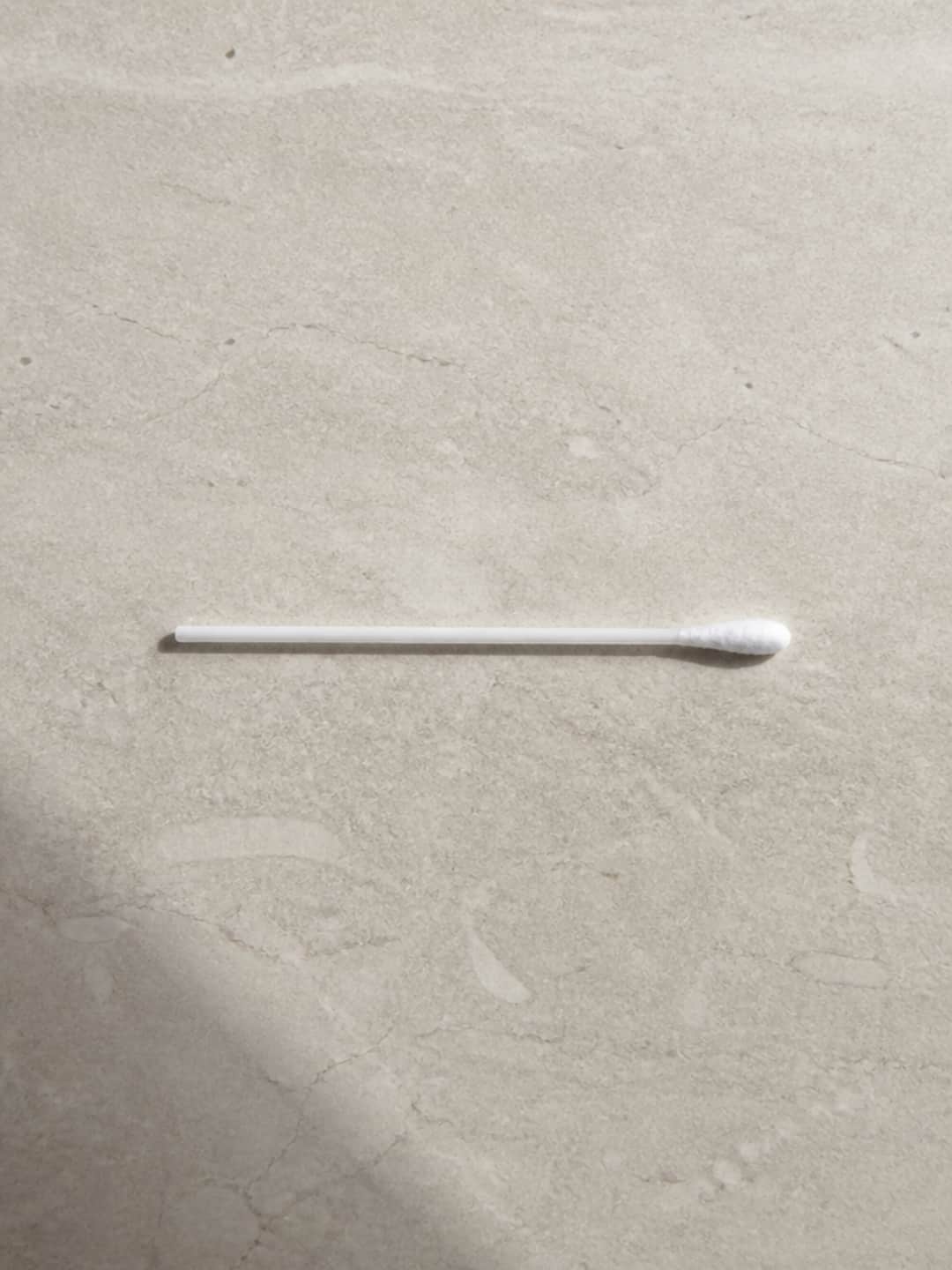 A single cotton swab lying on a pale warm-grey stone surface, shot from directly above in soft morning light