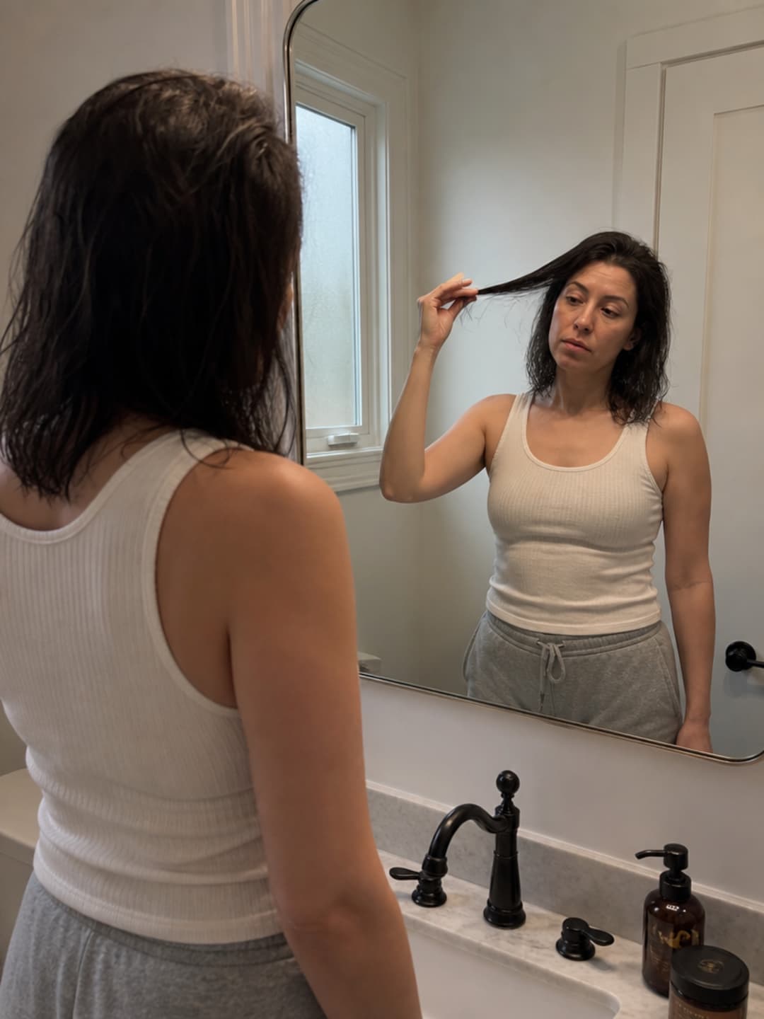 A woman of 34 with dark shoulder-length damp hair in a cream tank top stands at her bathroom mirror; her fingers are visibly stalled halfway down a section of hair she is testing — a flat, tired expression of recognition on her face