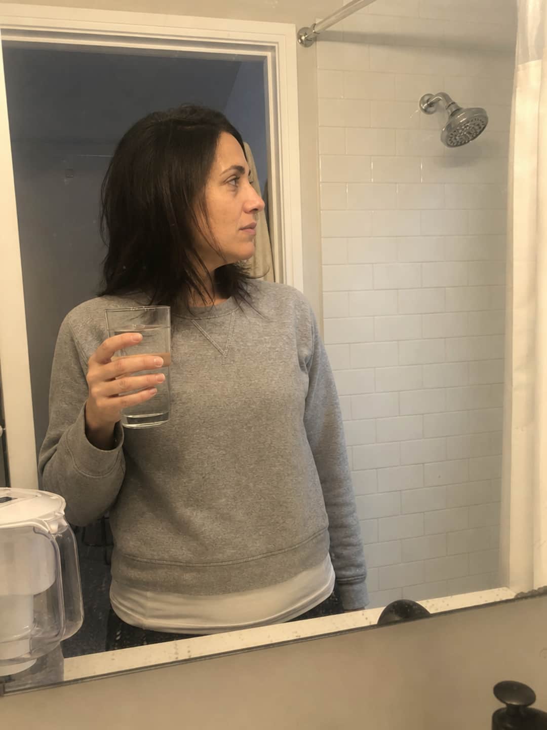 Maya, 38, stands at her bathroom counter holding a glass of tap water, looking toward the unfiltered chrome shower head visible behind her — a Brita pitcher at the edge of frame. The expression captures the moment of realizing the shower was the one water source she never filtered.