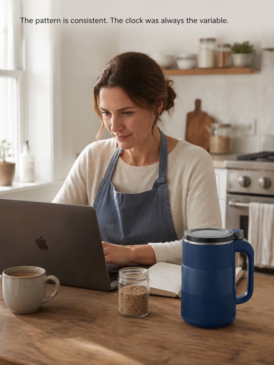 Woman seated at a kitchen table with a laptop, reading with focused engagement, a small clear container of wheat berries and the Grainbloom mill nearby, morning window light