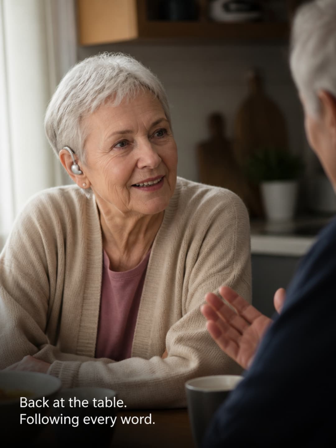 Person in their late 60s seated at a dining table following a conversation clearly, small behind-the-ear device visible