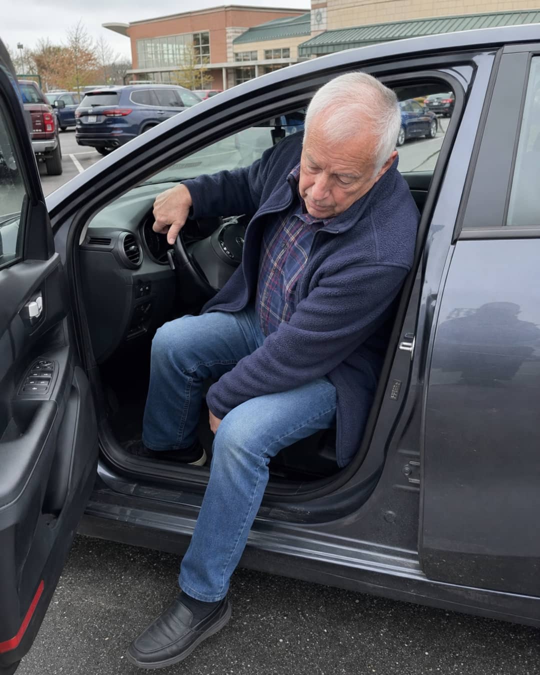 Man in his early 70s mid-exit from a sedan in a grocery store parking lot, right hand gripping the steering wheel at an awkward cross-body angle — the improvised grip that was never designed for this moment