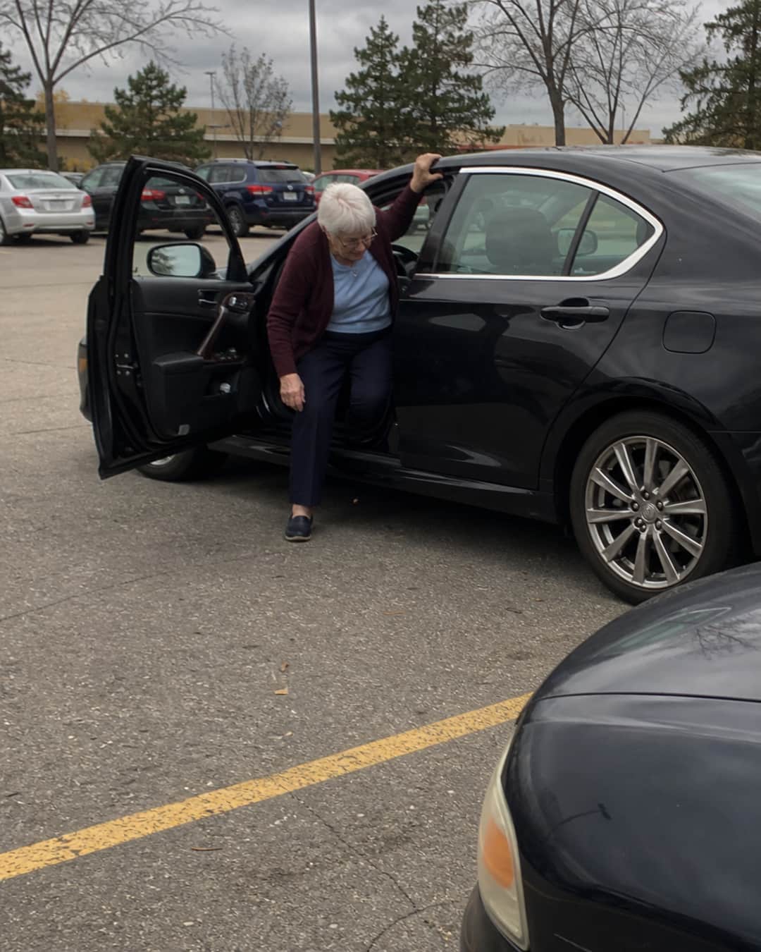 Candid parking lot photo: a woman of 71 with short silver-white hair in a burgundy cardigan is mid-exit from a dark sedan, her left hand gripping the smooth top edge of the door frame — body torqued, weight uneven, one foot on asphalt and one still inside the car, expression showing concentrated effort