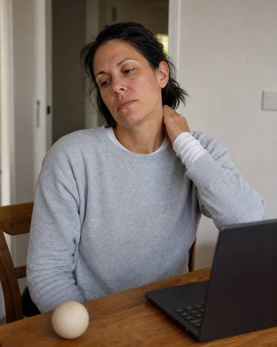 A woman of 38, dark shoulder-length hair pulled loosely back, wearing a soft heather-grey crewneck over a white long-sleeve shirt — sitting at a wooden desk with her thumb pressed into her neck–shoulder junction, expression flat with familiar frustration, a lacrosse ball unused beside her laptop.