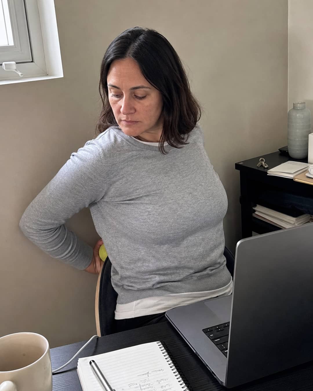 A woman of 34 with shoulder-length dark hair, in a light grey crewneck, seated at a home-office desk, arm twisted awkwardly behind her as she presses a lacrosse ball against the upper-back region — the specific flatness of someone performing a routine she has done many times