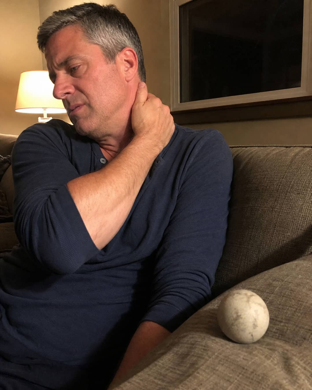 A man of 47 with short salt-and-pepper hair wearing a faded navy henley, seated on a couch in a dimly lit living room, pressing his left thumb firmly into the right side of his neck where it meets the upper trapezius — a lacrosse ball visible on the cushion beside him, his expression caught between effort and frustration