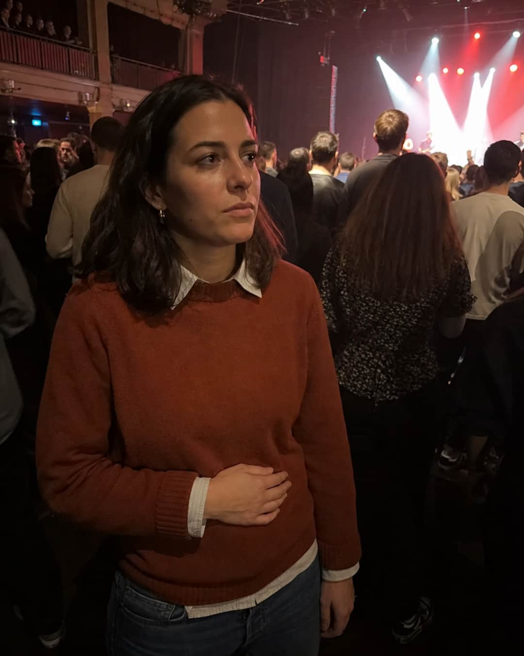 A woman of 28 with dark shoulder-length hair in a rust-colored sweater stands inside a dimly lit music venue; one hand presses flat and low across her abdomen, weight shifted to one hip, eyes focused in the middle distance while the crowd and stage lights continue behind her — present at the concert but pulled slightly outside the moment