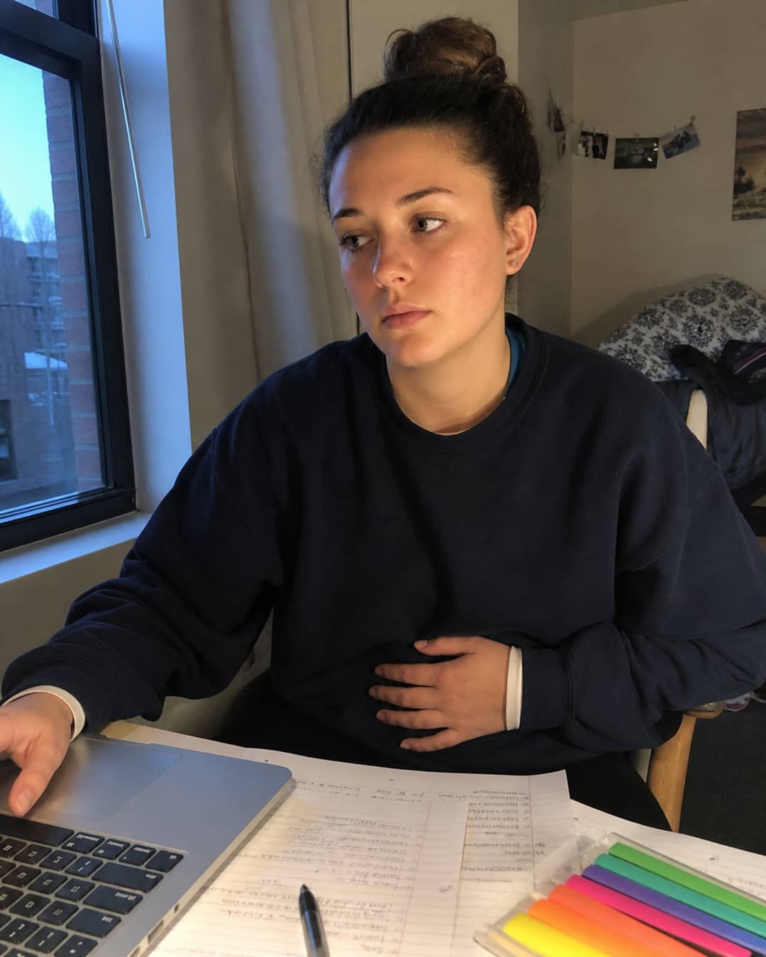 Candid photo of a 24-year-old woman with brown hair in a high bun, wearing a navy crewneck sweatshirt, seated at a study desk late at night with open notes and a laptop, one hand pressed lightly against her lower abdomen, expression turned away — the posture of distraction, not the work